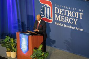President Garibaldi stands at a podium inside of the Student Fitness Center, with two blue University of Detroit Mercy Build A Boundless Future banners, one on the podium and one as a large backdrop behind him.