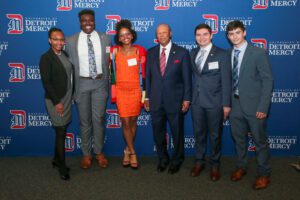 Six people stand inside of the Student Fitness Center with a blue University of Detroit Mercy backdrop behind them.
