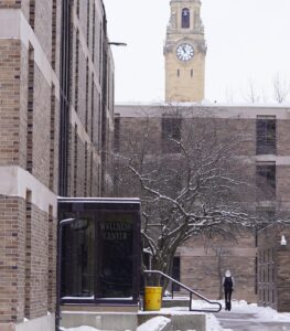 An outdoor photo of the Quads dorms with the Wellness Center in the foreground, a student walking on a snowy sidewalk and the clock tower looming beyond the dorms.