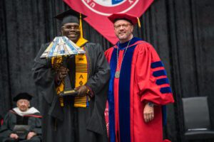 A student holds a lamp on a stage, wearing graduation attire, standing next to President Donald Taylor, wearing red Commencement robes.