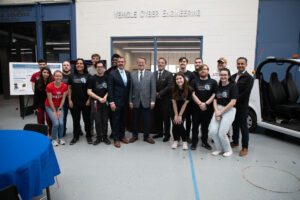 More than a dozen students and others pose with Gary Peters and Harry Coker Jr. inside of the Engineering Building High Bay, with Vehicle Cyber Engineering on the wall above.