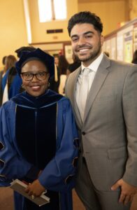 Two people stand indoors smiling, one wearing graduation gown and cap and the other wearing a suit.