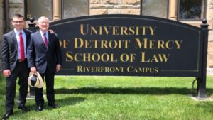 Two people stand outdoors next to University of Detroit Mercy School of Law, Riverfront Campus sign.