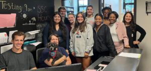 Eleven people stand and pose for a photo inside of the Student Success Center, two of them sitting and one holding a dog.