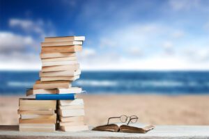 Books are stacked on a beach with water in the background.