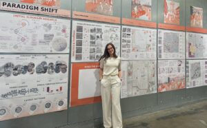 Iryna Olkhovetska stands in front of a series of designs and pictures located on the wall inside of the School of Architecture + Community Development.