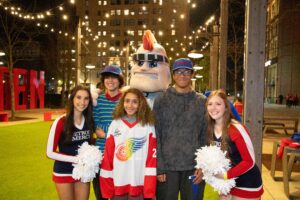 Five people stand outdoors during Detroit Mercy Night at Little Caesars Arena, with Tommy Titan also in the photo.