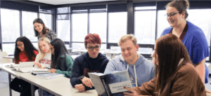 A photo of students at desks with textbooks open studying and interacting with instructors.