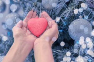 Female hands holding a heart on the blue background of a Christmas tree.