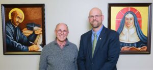 Two leaders at the Law school stand next to portraits of St. Ignatius of Loyola and Catherine McAuley.