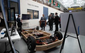 A photo of four men talking inside of the high bay of the Engineering Building, with a model car next to them.