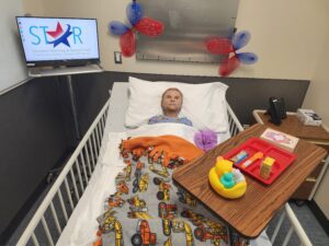 The new STAR Center child-size mannequin lays in a hospital bed, under a blanket with images of tractors and trucks on it, with tray table of kids toys and food close to the bed. The STAR Center logo is displayed on the tv behind the mannequin.