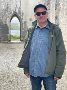 An image of a man wearing sunglasses standing in front of a stone wall with a peaked doorway.