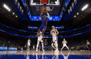 An image of a Detroit Pistons player going up for a dunk shot in Little Caesar's Arean with Sacramento King players watch in the background