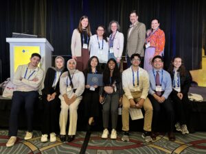 Thirteen people stand and sit and pose for a photo inside of a conference room near a stage.