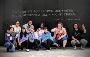 An image of several people crouched under words on a black wall. The words are a quote from Martin Luther King, Jr., "...until justice rolls down like waters and righteousness like a mighty stream."