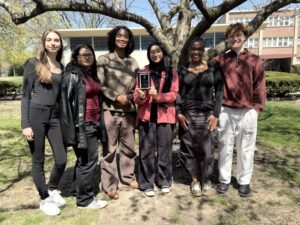 Six students stand outdoors underneath a tree on the McNichols Campus during a sunny day.