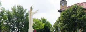 A summer outdoor photo of the McNichols Campus featuring the Jesus statue, trees, buildings and the clock tower.
