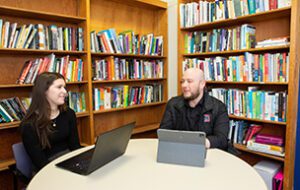 Two students talk while working next to bookshelves.
