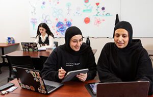 Two students work on their laptops in the foreground, while behind them a student works on her laptop.