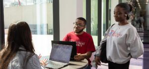 Three students gather around a table on their laptops, while one student stands.