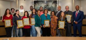 Fifteen people receive awards and stand and pose for a photo inside of a boardroom.