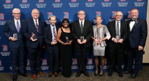 Group of eight individuals standing in front of a blue backdrop with repeating “Detroit Mercy” logos. Each person is dressed in formal attire and holding a glass award.