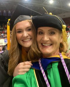 Two women in graduation attire smile for a photo inside of Calihan Hall during a commencement ceremony.