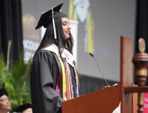 Student speaker at 2025 UDM Commencement at the podium on stage with colorful flowers in the foreground