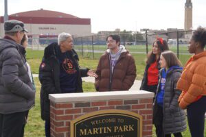 Seven people stand outdoors near a sign for Martin Park and the perimeter of the McNichols Campus.