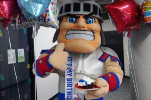 An image of UDM's Tommy Titan standing under balloons holding a piece of cake in one hand and the other hand with thumb's up sign.
