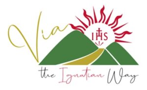 Logo for Via. The word “Via” appears in gold script above a graphic of three green hills. A red Jesuit IHS emblem with radiating sun rays rises behind the hills, and a gold path winds between them. Beneath the hills, the text reads “The Ignatian Way.”