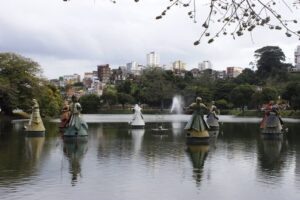 Decorative statues standing in a calm lake with a fountain at the center, surrounded by trees and a city skyline in the background under an overcast sky.