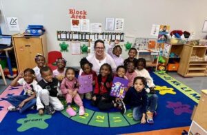 A teacher sits with a group of young children on a colorful alphabet rug in an early childhood classroom. Several children hold decorated purple craft projects, while shelves, learning materials and a wall display labeled are visible in the background.