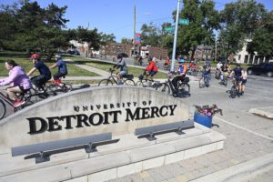 People ride bikes past the University of Detroit Mercy sign near the main entrance during Homecoming on a sunny, fall day.