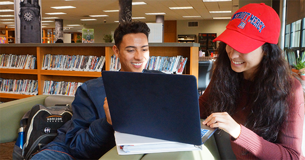 two students on a laptop in the library. one is wearing a detroit mercy hat.