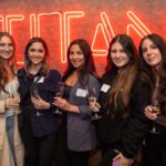 Five students hold champagne glasses and smile in front of a bright TITAN sign inside of the Titan Club.