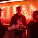 Three students clink champagne glasses in front of the large TITAN sign inside of the Titan Club.