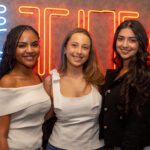 Three students smile and pose for a photo inside of the Titan Club, with champagne glasses and a brightly lit TITAN sign in the background.