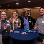 Four students with champagne glasses smile and pose for a photo around a table inside of the Titan Club.